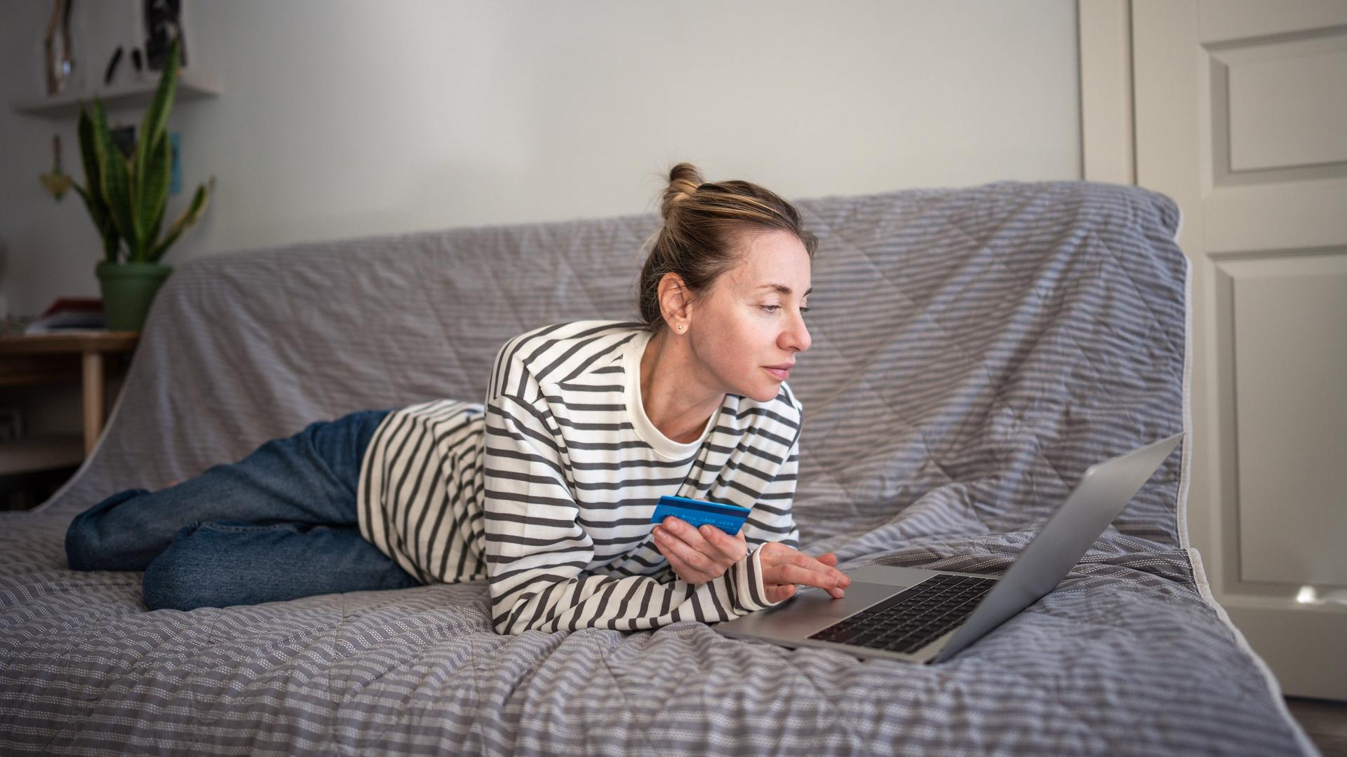 Eine Frau, mit einer Bankkarte in der Hand, liegt auf der Couch und benutzt einen Laptop