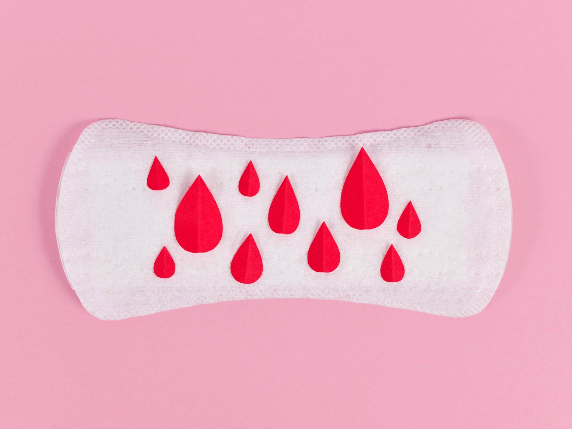 Aufnahme einer Slipeinlage auf rosa Untergrund. Auf der Einlage sind kleine rote Blutstropfen aus Papier verteilt.