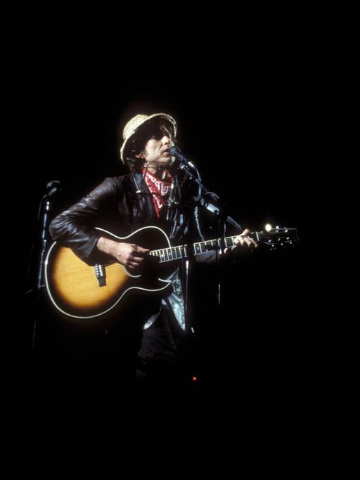 Bob Dylan steht mit Gitarre  in einem hellen Spot auf einer sonst dunklen Bühne während eines Konzertes 1984 in München.