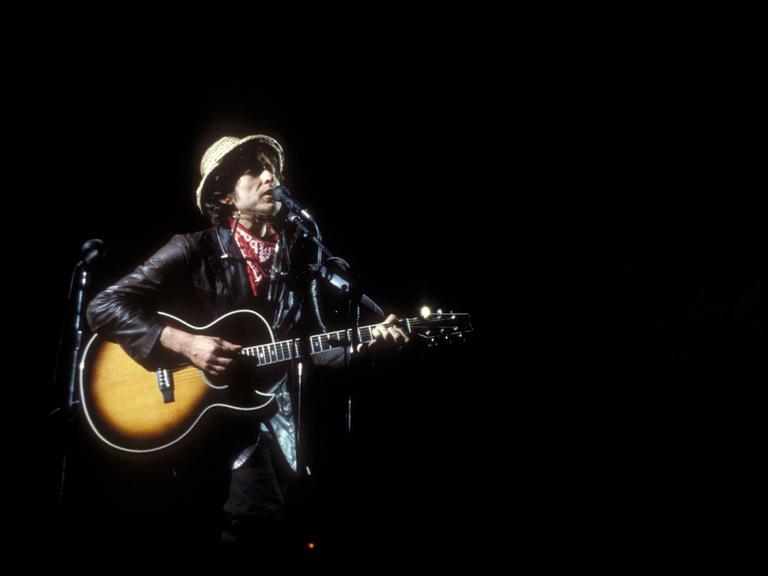 Bob Dylan steht mit Gitarre  in einem hellen Spot auf einer sonst dunklen Bühne während eines Konzertes 1984 in München.