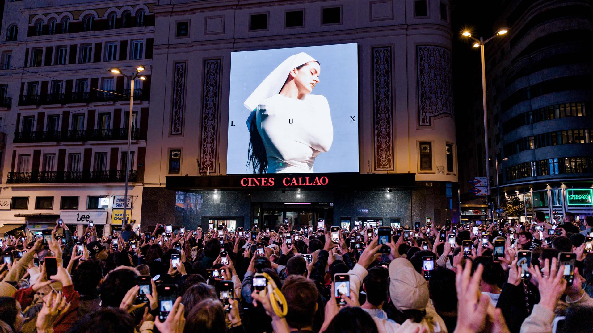 Eine auf einem Platz versammelte Menschenmenge fotografiert mit Smartphones das Cover von Rosalías aktuellem Album.