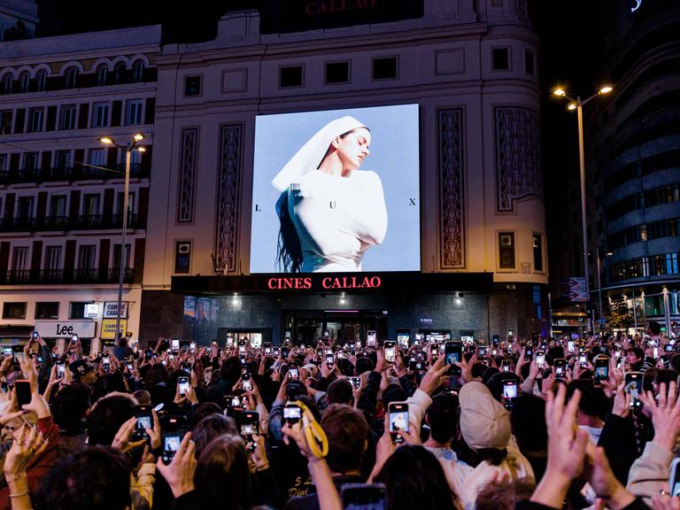 Eine auf einem Platz versammelte Menschenmenge fotografiert mit Smartphones das Cover von Rosalías aktuellem Album.