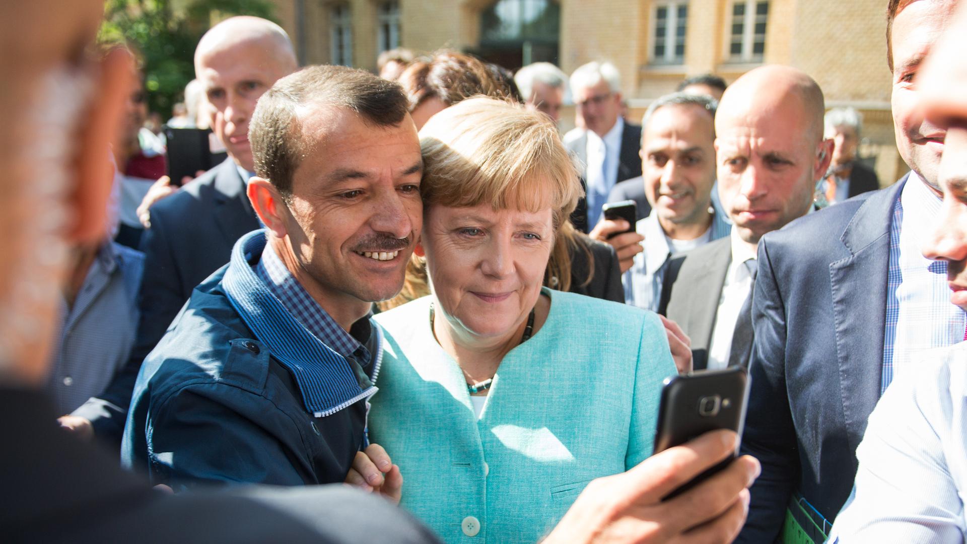 Die beiden stehen vor einem Haus Wange an Wange nebeneinander; Merkel trägt ein türkisfarbenes Kostüm. Dahinter zahlreiche weitere Personen. Die beiden stehen vor einem Haus Wange an Wange nebeneinander; Merkel trägt ein türkisfarbenes Kostüm. Dahinter zahlreiche weitere Personen.