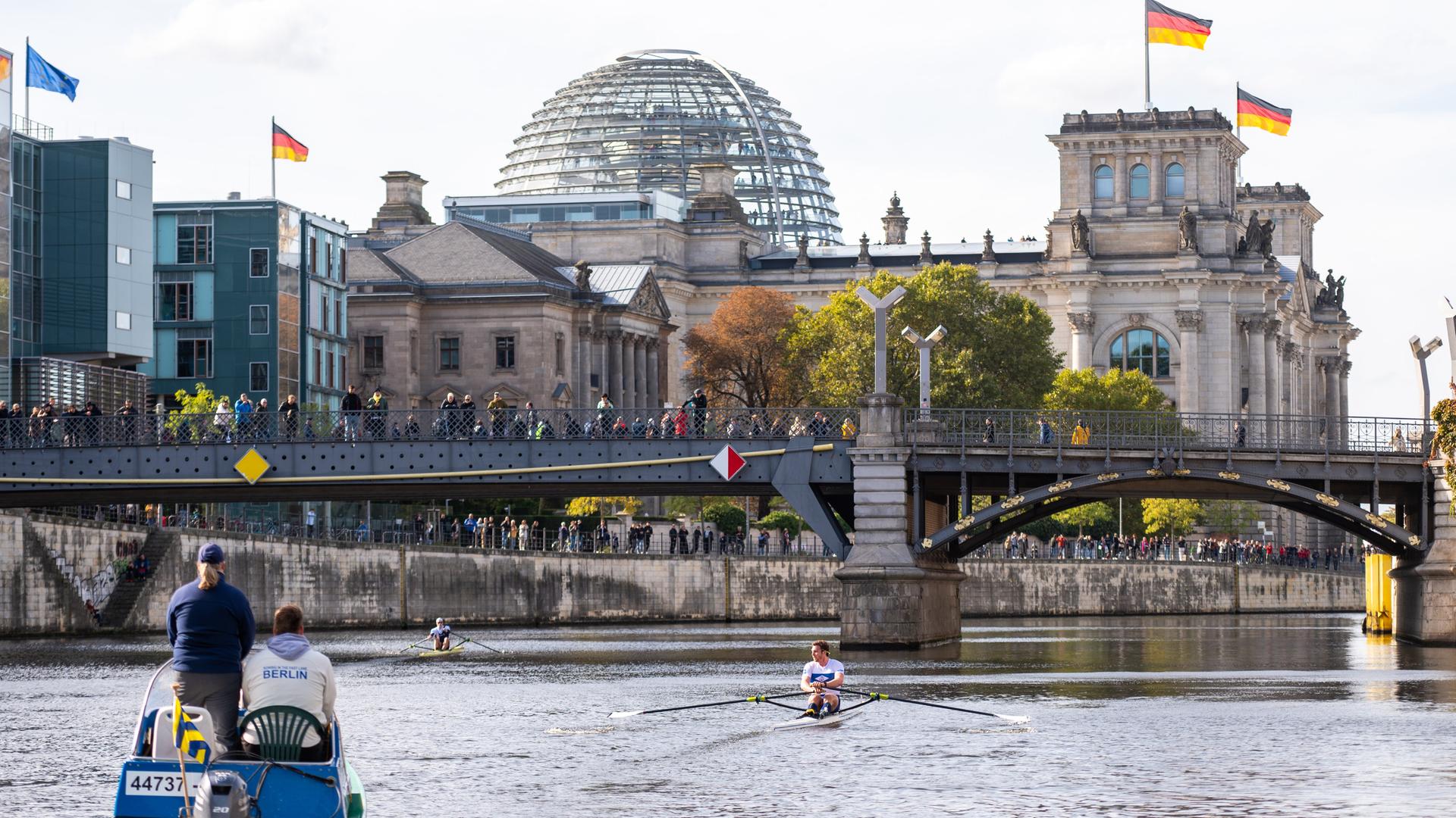 2 Ruder-Boote auf dem Fluss Spree in Berlin. Auf einer Brücke stehen Menschen und schauen den Ruderern zu. Im Hintergrund ist der Reichs-Tag zu sehen.