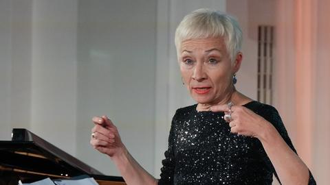 Eine Frau mit kurzem weißblonden Haar performt vor einem Flügel. Sie gestikuliert mit den Fingern und trägt ein schwarzes, glitzerndes Kleid.