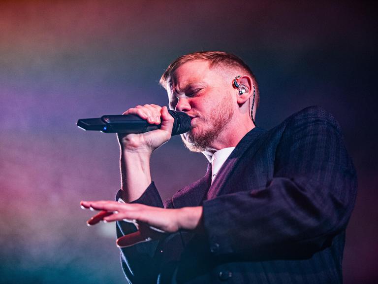 Kae Tempest bei einem Auftritt am 26.10.2025 in Bologna. Kae Tempest trägt einen Anzug, rappt in ein Mikro und gestikuliert mit der anderen Hand.