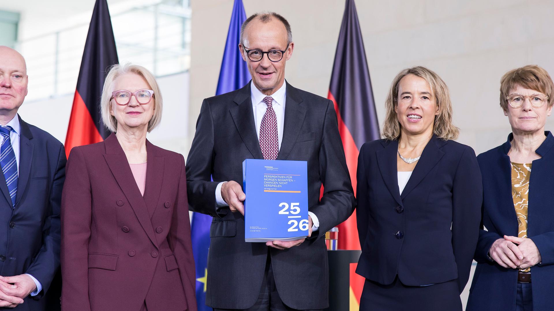 Prof. Dr. Monika Schnitzer, Friedrich Merz und Mitglieder des Sachverständigenrates stehen dicht nebeneinander bei der Übergabe des Jahresgutachtens 2025/2026.