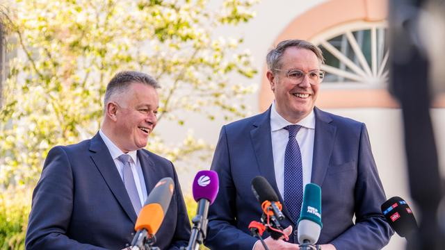 Zwei Männer in Anzügen stehen nebeneinander vor vielen Mikrofonen, beide lächeln.