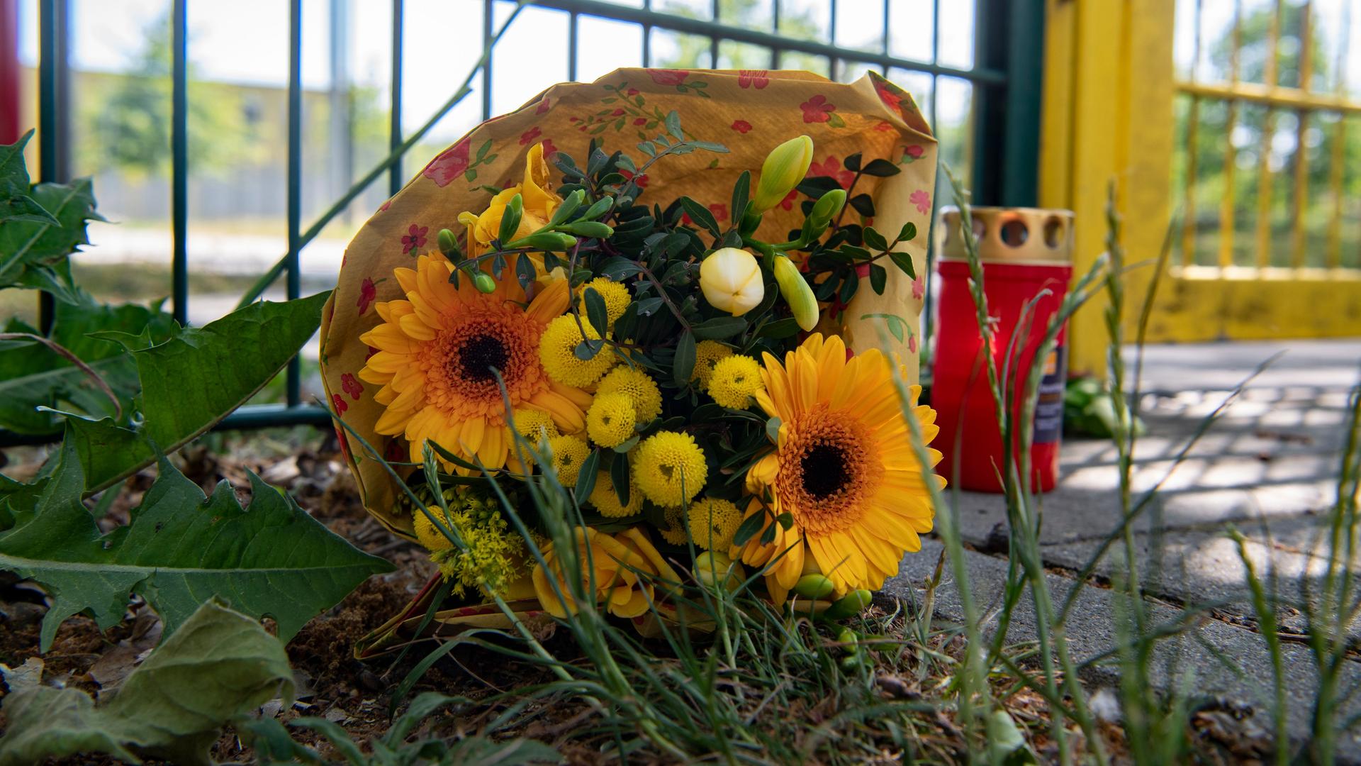 Blumen mit gelben Farbtönen und eine rotleuchtende Kerze liegen neben einem Zaun. Blumen mit gelben Farbtönen und eine rotleuchtende Kerze liegen neben einem Zaun.