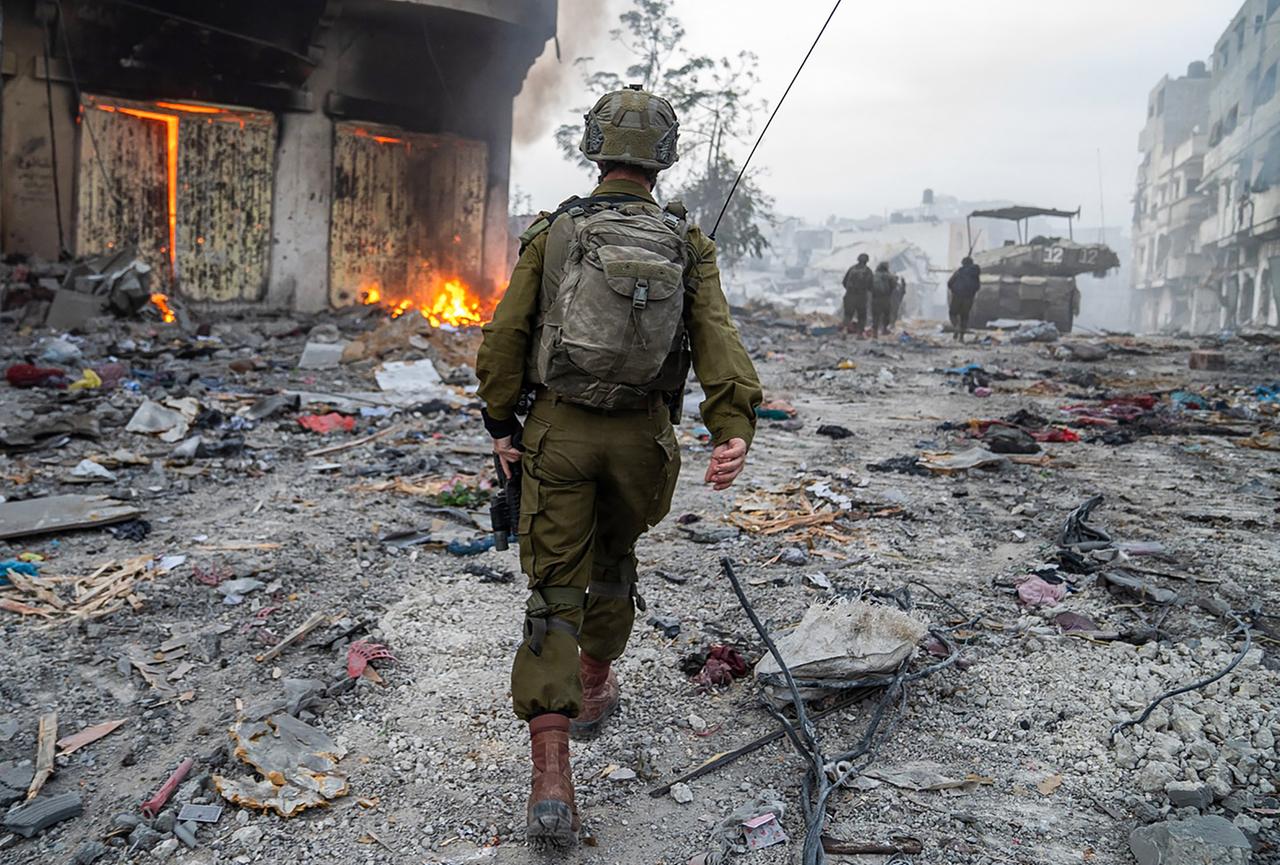 Ein israelischer Soldat geht über einen mit Trümmern übersäten Platz neben einem brennenden Haus.