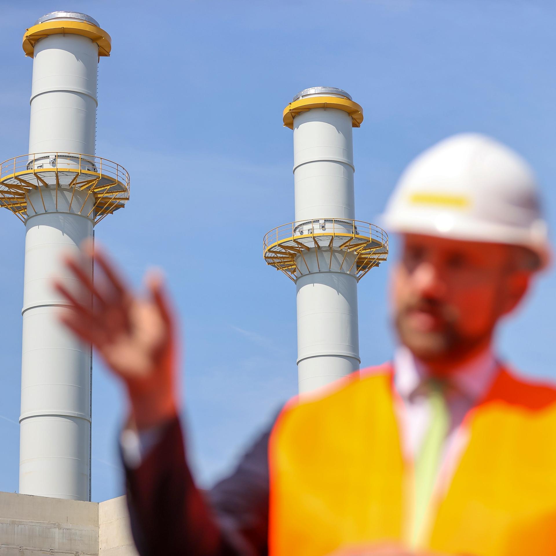Eine Person mit Schutzhelm und Warnweste hebt sich auf einer Baustelle unscharf vor Fabrikschloten ab.