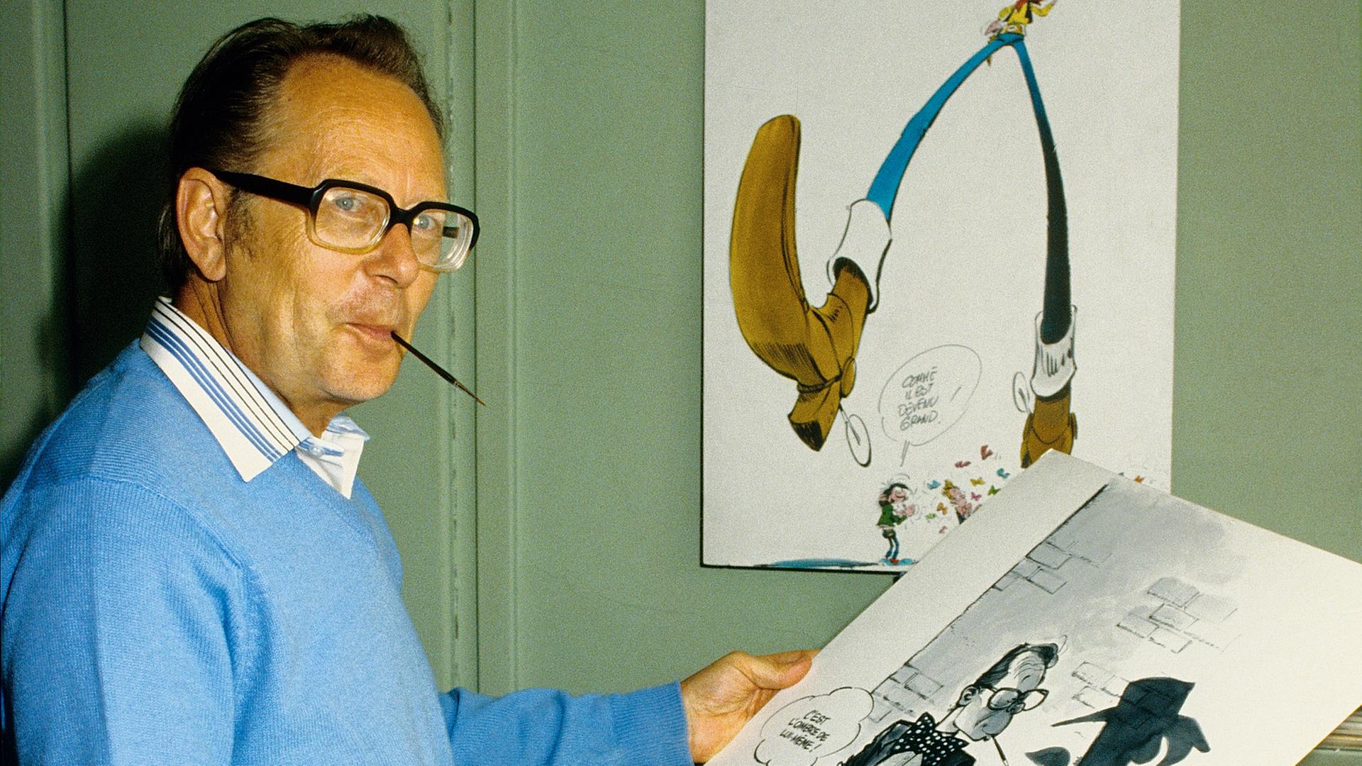 Der Zeichner Maurice De Bevere (Morris), Erfinder von Lucky Luke, steht mit einer großen Skizze in der Hand und einem Zeichenstift im Mund in seinem Büro (1984).