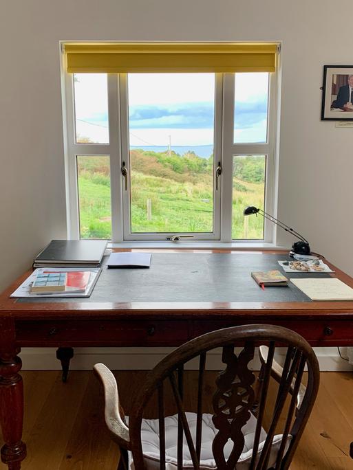 Ein Schreibtisch am Fenster mit Blick auf eine grüne Wiese und das Meer