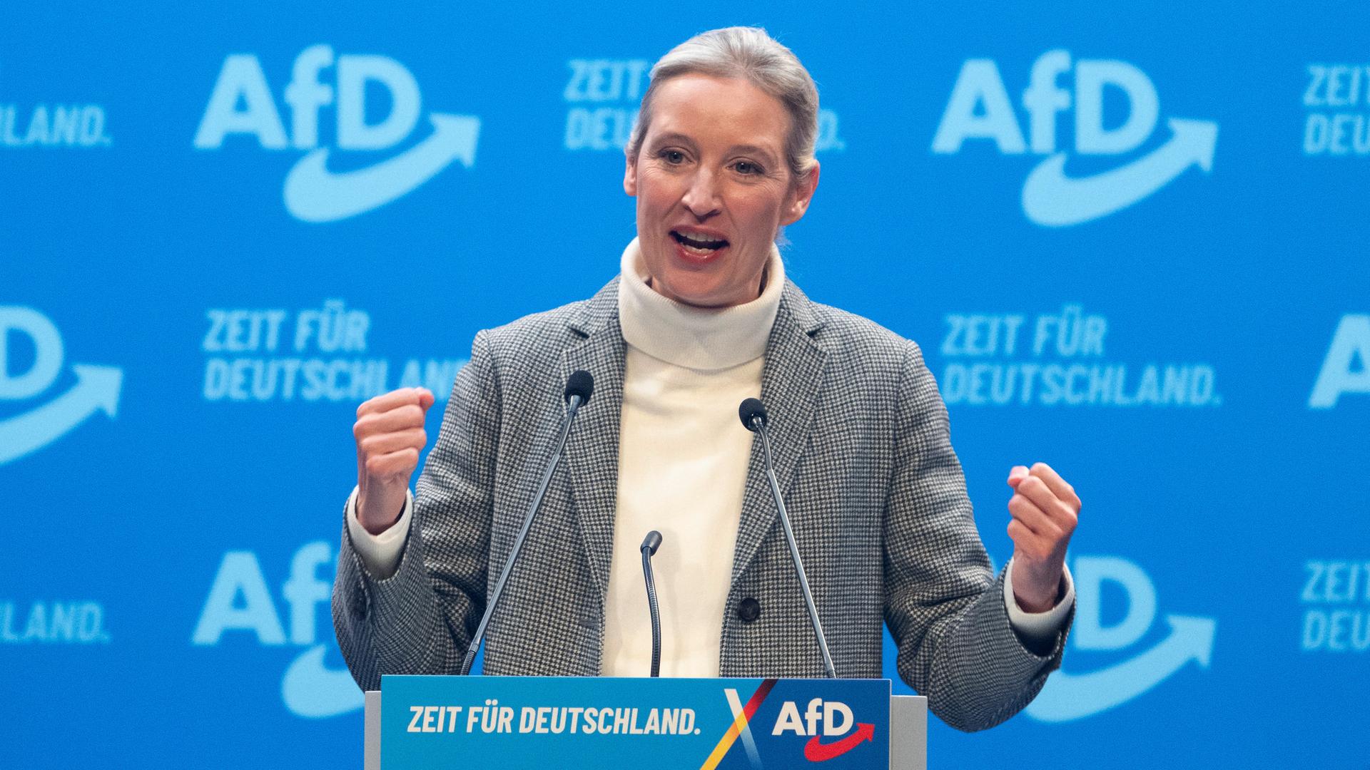 Alice Weidel steht an einem Rednerpult und hat beide Hände zur Faust geballt. Im Hintergrund eine Wand mit dem AfD-Logo.