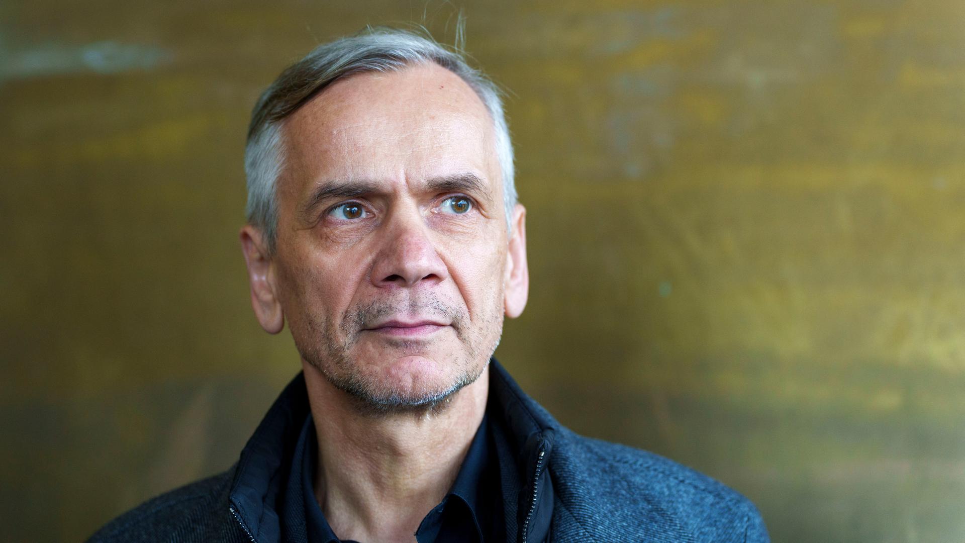 Lutz Seiler blickt ernst ins Off des Bildes. Der Bildhintergrund ist bronzefarben