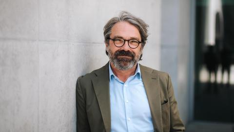 Portrait eines Mannes mit graumelierten Haaren und einer schwarzen Brille, der an einer Betonwand lehnt und in die Kamera lächelt