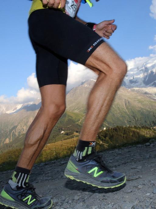 Nahaufnahme der Beine eines Läufers in professioneller Ausrüstung, der einen Pfad in den Alpen bergauf läuft.