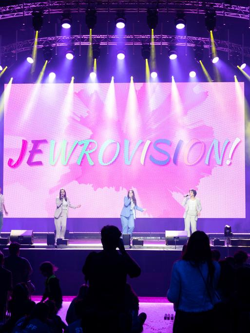 Zu sehen ist eine Bühne mit jungen Künstlerinnen und Künstlern. im Hintergrund das Logo von dem Wettbewerb Jewrovision.