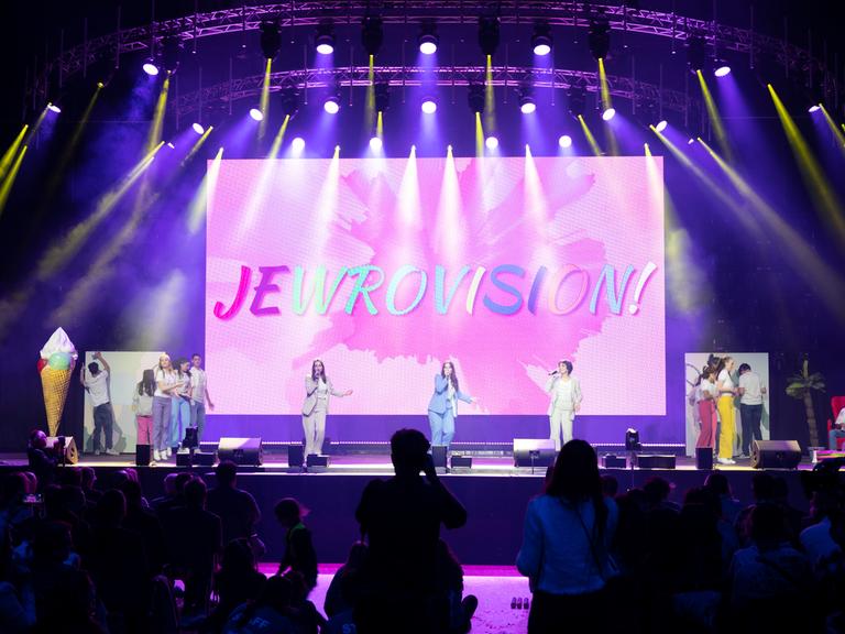 Zu sehen ist eine Bühne mit jungen Künstlerinnen und Künstlern. im Hintergrund das Logo von dem Wettbewerb Jewrovision.
