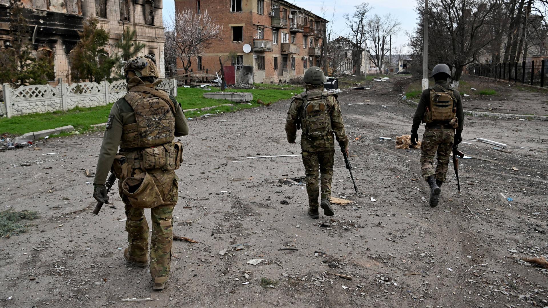 Drei bewaffnete Männer in Soldatenuniformen patrouillieren durch eine vom Krieg schwer beschädigte Kleinstadt.