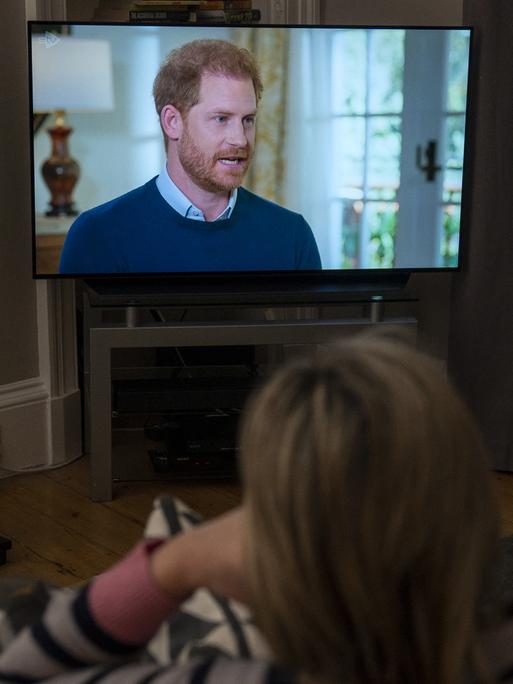 Eine Frau sitzt im Wohnzimmer vor dem Fernseher und schaut das Interview mit dem britischen Prinzen Harry an. 