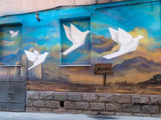 Ein Wandgemälde an einer Hausfassade zeigt mehrere weiße Tauben, die gen Himmel fliegen. Im Boden steckt ein Schild auf dem "Minen" steht. 