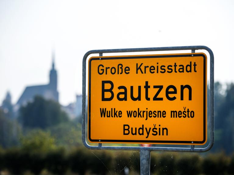 Ein Ortsschild der Stadt Bautzen, im Hintergrund unscharf ein Kirchturm.
