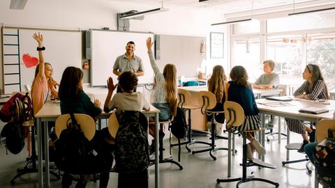 Schüler sitzen in einem Klassenraum. Vorn steht ein Mann mit dunklen, kurzen Haaren, der die Gruppe anlacht.