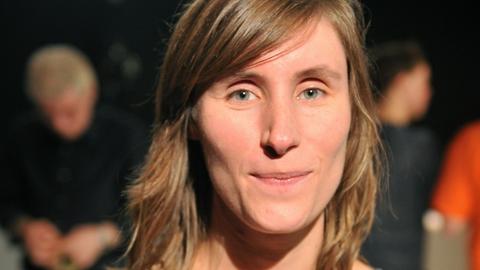 Julia Roesler trägt ein graues Oberteil und schaut freundlich und direkt in die Kamera. Im Hintergrund sind in der Unschärfe weitere Menschen zu sehen.
