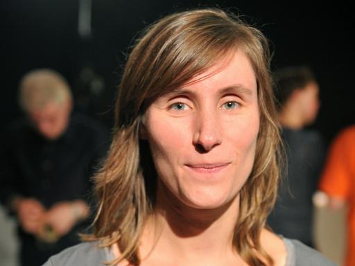 Julia Roesler trägt ein graues Oberteil und schaut freundlich und direkt in die Kamera. Im Hintergrund sind in der Unschärfe weitere Menschen zu sehen.