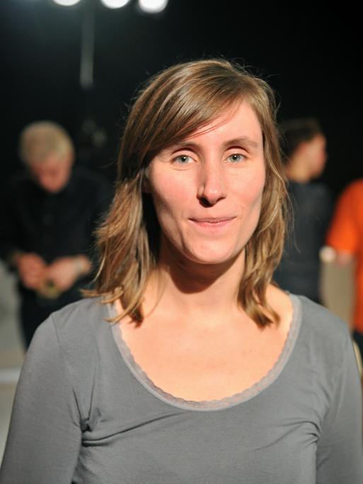 Julia Roesler trägt ein graues Oberteil und schaut freundlich und direkt in die Kamera. Im Hintergrund sind in der Unschärfe weitere Menschen zu sehen.