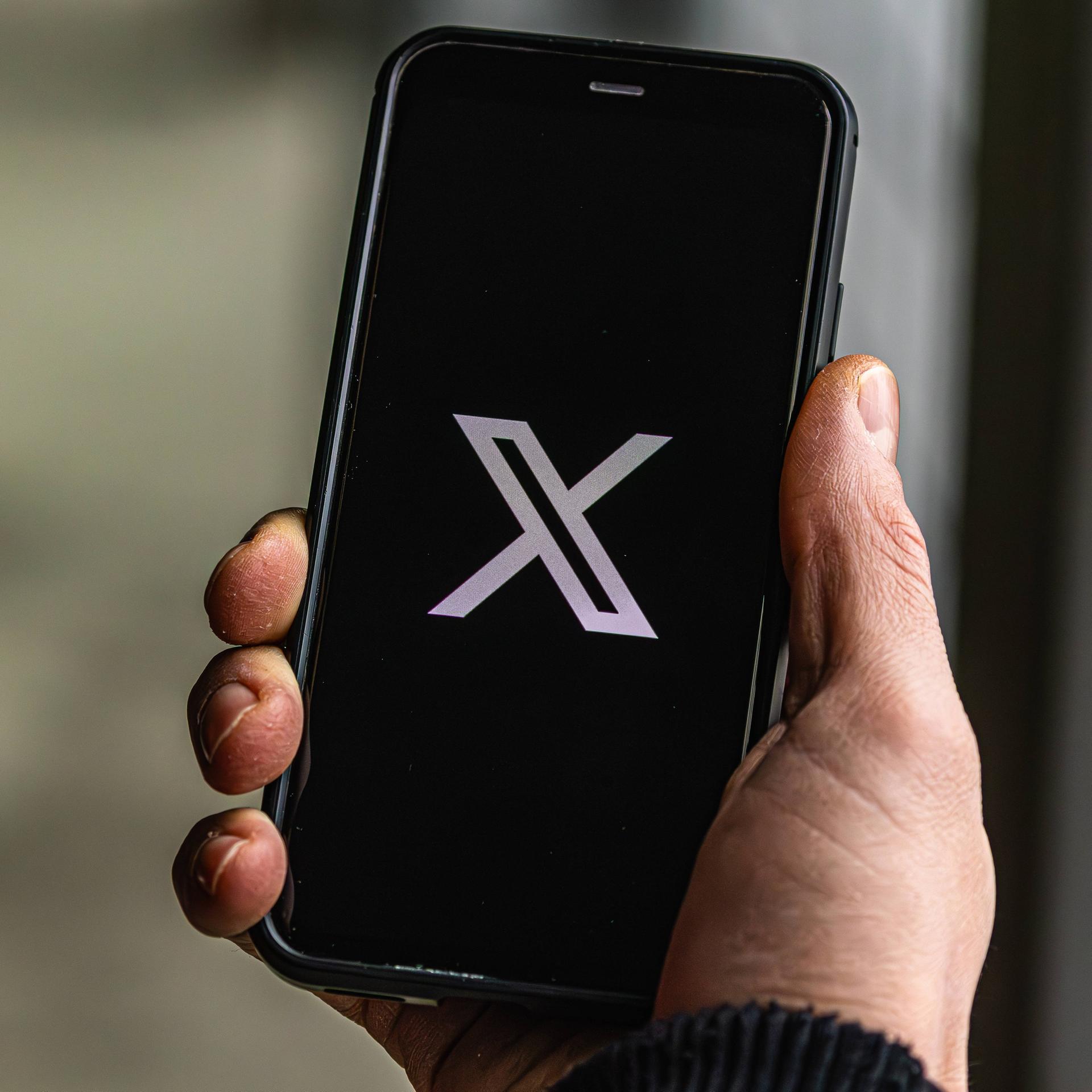 Jemand hält ein Handy in der Hand auf dem die Plattform "X" geöffnet wird.