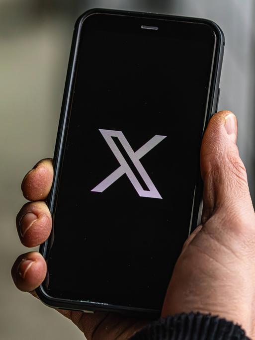 Jemand hält ein Handy in der Hand auf dem die Plattform "X" geöffnet wird.