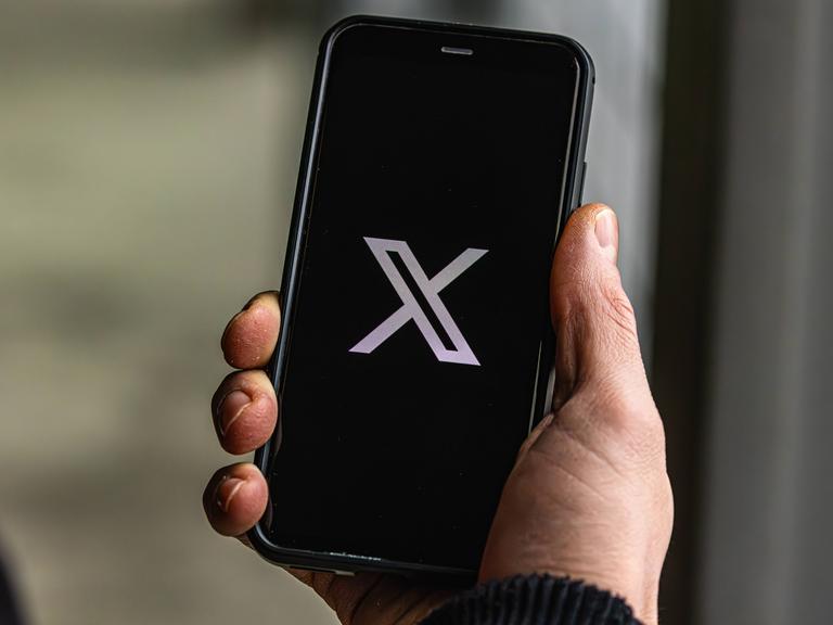 Jemand hält ein Handy in der Hand auf dem die Plattform "X" geöffnet wird.