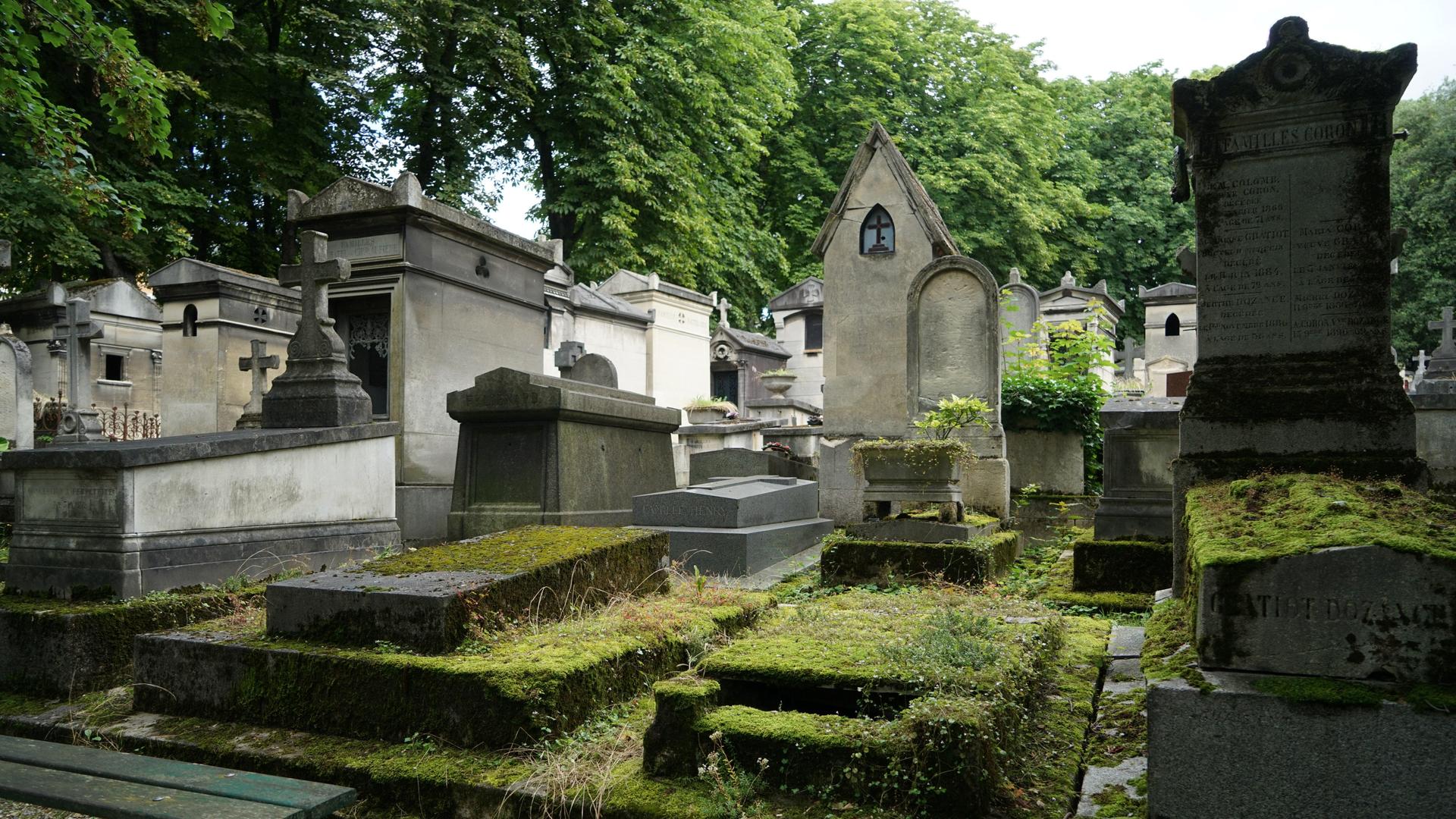 Das Bild zeigt mehrere Gräber und Gruften auf dem Pariser Friedhof Père-Lachaise, die teilweise stark mit Moos bewachsen sind. 