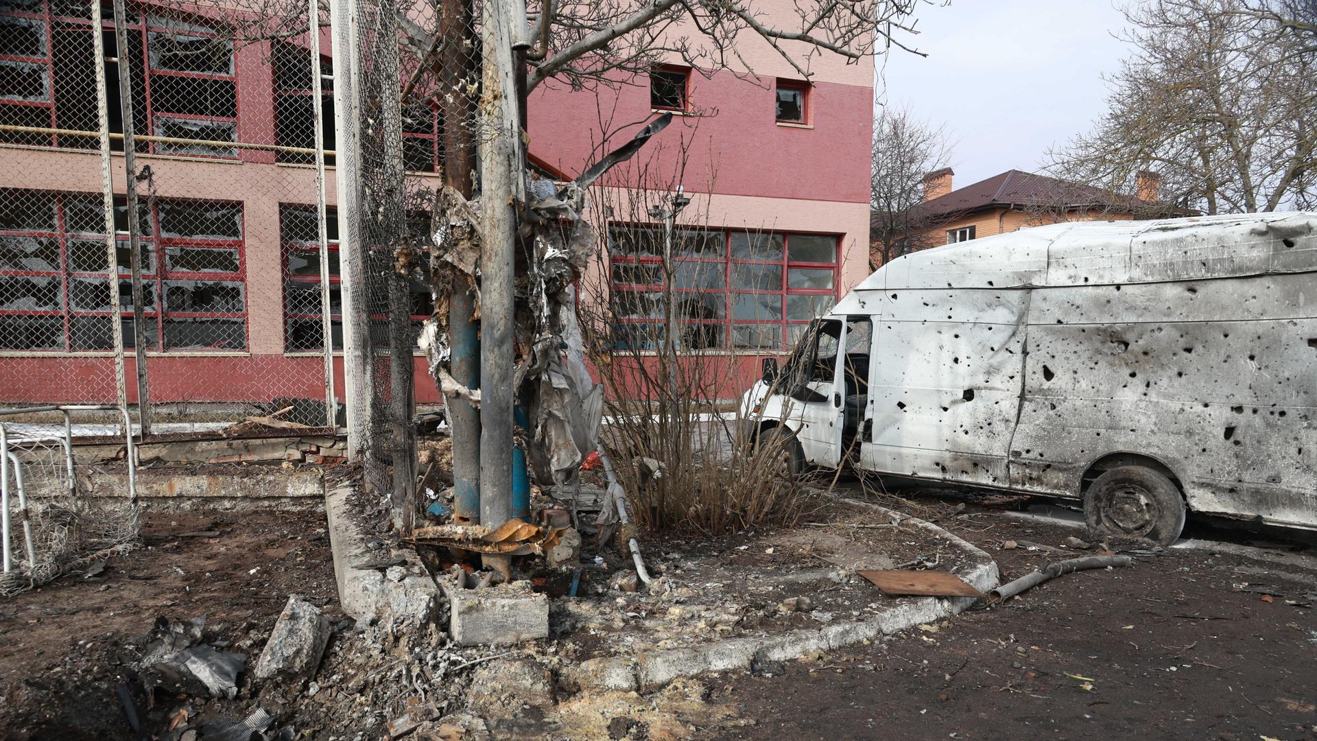 Vor einem Gebäude mit zerbrochenen Fensterscheiben steht ein von Metallgeschossen durchlöcherter Transporter.