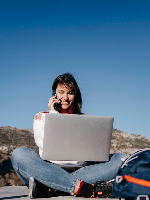 Arbeiten, wo andere Urlaub machen - für viele ein Traum. Zu sehen: Eine junge Frau sitzt auf einem Plateau (im Hintergrund sind Berge), hat einen Laptop auf dem Schoß und telefoniert mit einem Handy. 