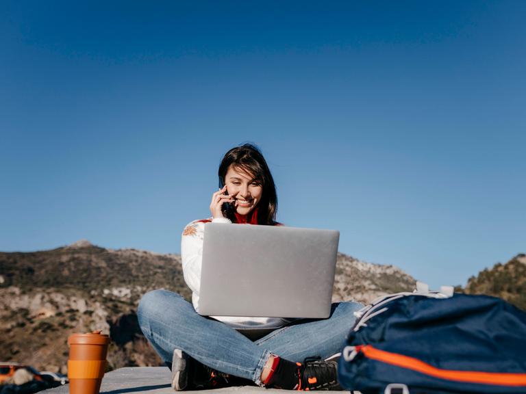 Arbeiten, wo andere Urlaub machen - für viele ein Traum. Zu sehen: Eine junge Frau sitzt auf einem Plateau (im Hintergrund sind Berge), hat einen Laptop auf dem Schoß und telefoniert mit einem Handy. 