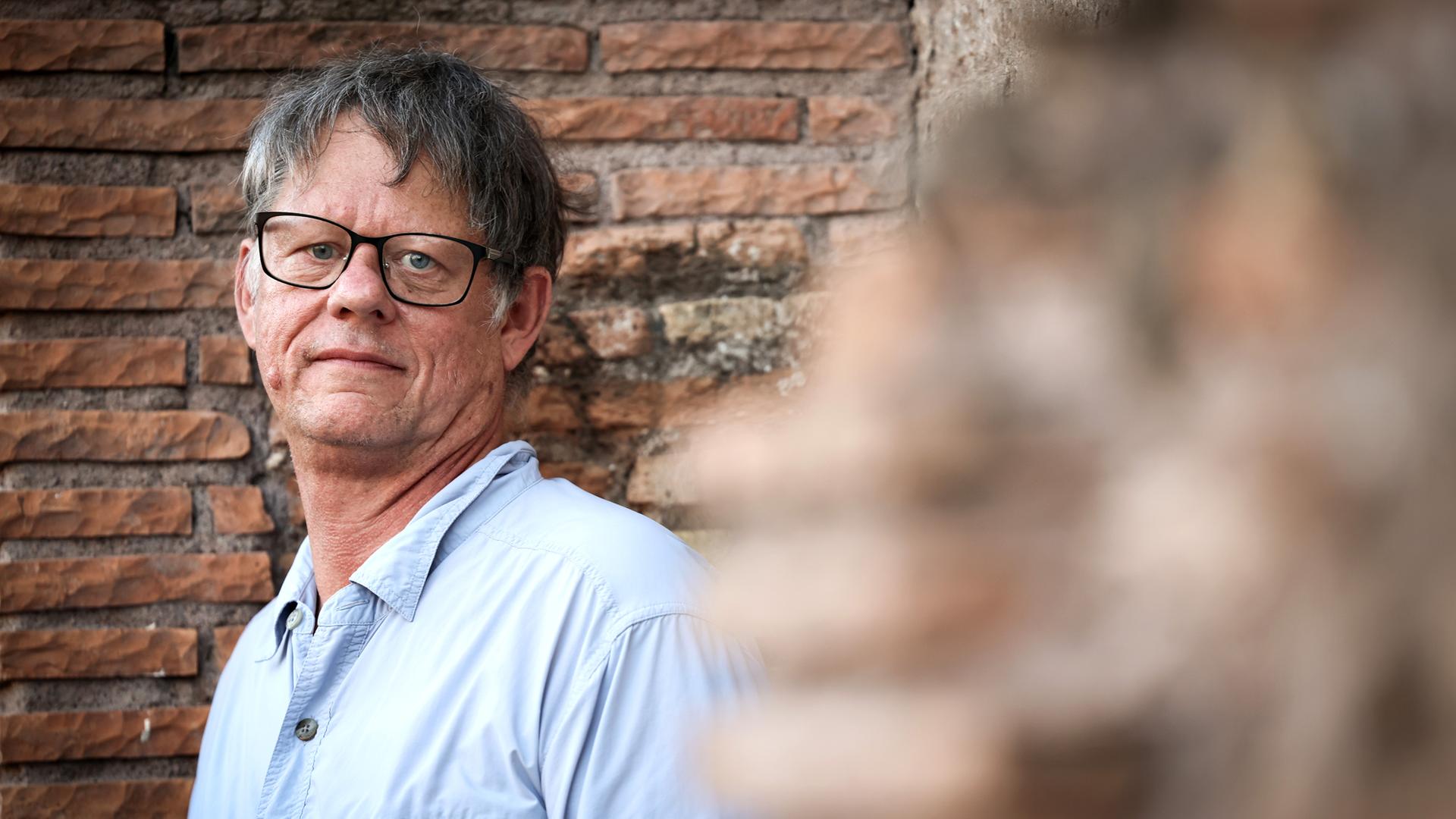 William T. Vollmann steht vor einer gemauerten Wand und schaut direkt in die Kamera. Er trägt ein helles Hemd und eine dunkle Brille. William T. Vollmann steht vor einer gemauerten Wand und schaut direkt in die Kamera. Er trägt ein helles Hemd und eine dunkle Brille.