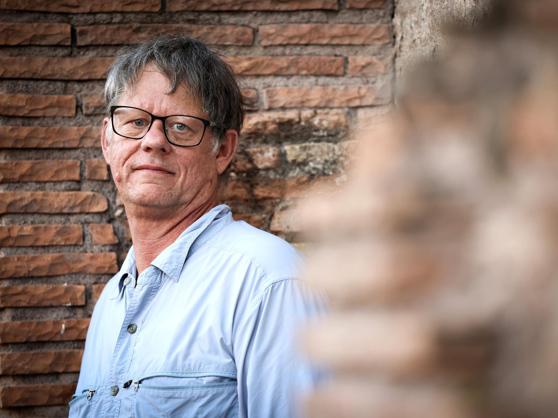 William T. Vollmann steht vor einer gemauerten Wand und schaut direkt in die Kamera. Er trägt ein helles Hemd und eine dunkle Brille.
