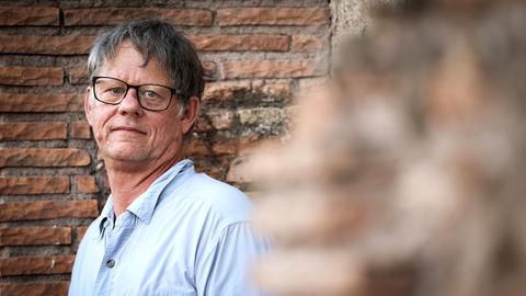 William T. Vollmann steht vor einer gemauerten Wand und schaut direkt in die Kamera. Er trägt ein helles Hemd und eine dunkle Brille. William T. Vollmann steht vor einer gemauerten Wand und schaut direkt in die Kamera. Er trägt ein helles Hemd und eine dunkle Brille.