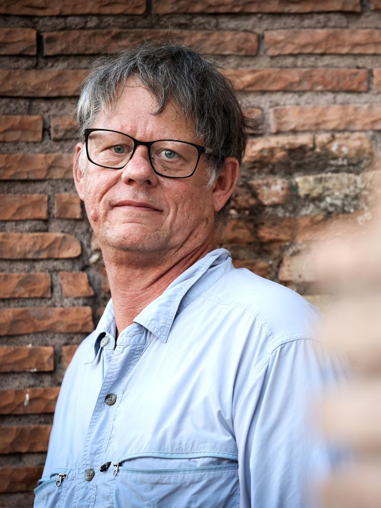 William T. Vollmann steht vor einer gemauerten Wand und schaut direkt in die Kamera. Er trägt ein helles Hemd und eine dunkle Brille.