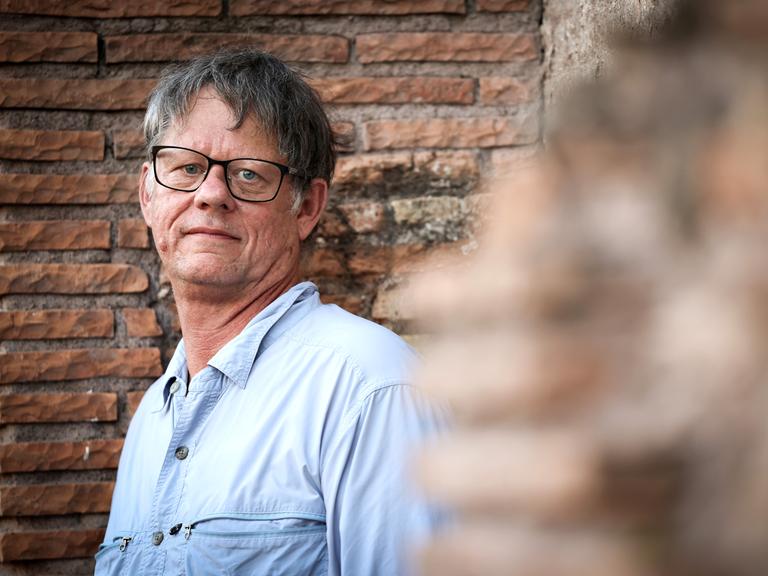 William T. Vollmann steht vor einer gemauerten Wand und schaut direkt in die Kamera. Er trägt ein helles Hemd und eine dunkle Brille.