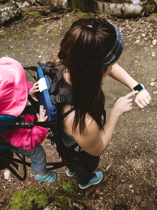 Eine junge Mutter überprüft die Zeit auf ihrer Smartwatch, während sie ihr Baby im Rucksack trägt.