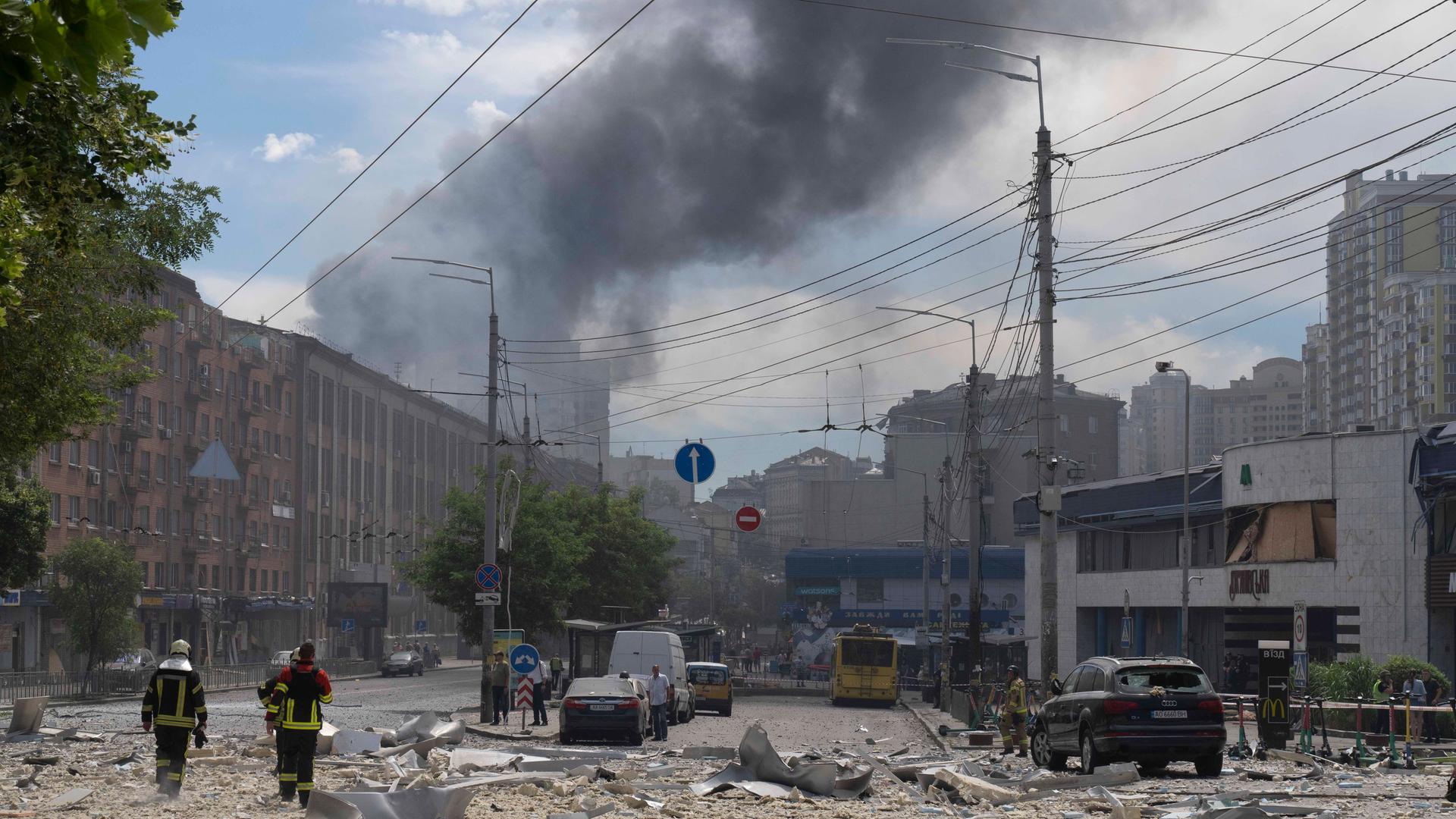 Rauch steigt nach einem russischen Angriff über einem Gebäude in Kiew auf, auf der Straße liegen Trümmer, zwei Feuerwehrmänner sind von hinten zu sehen.