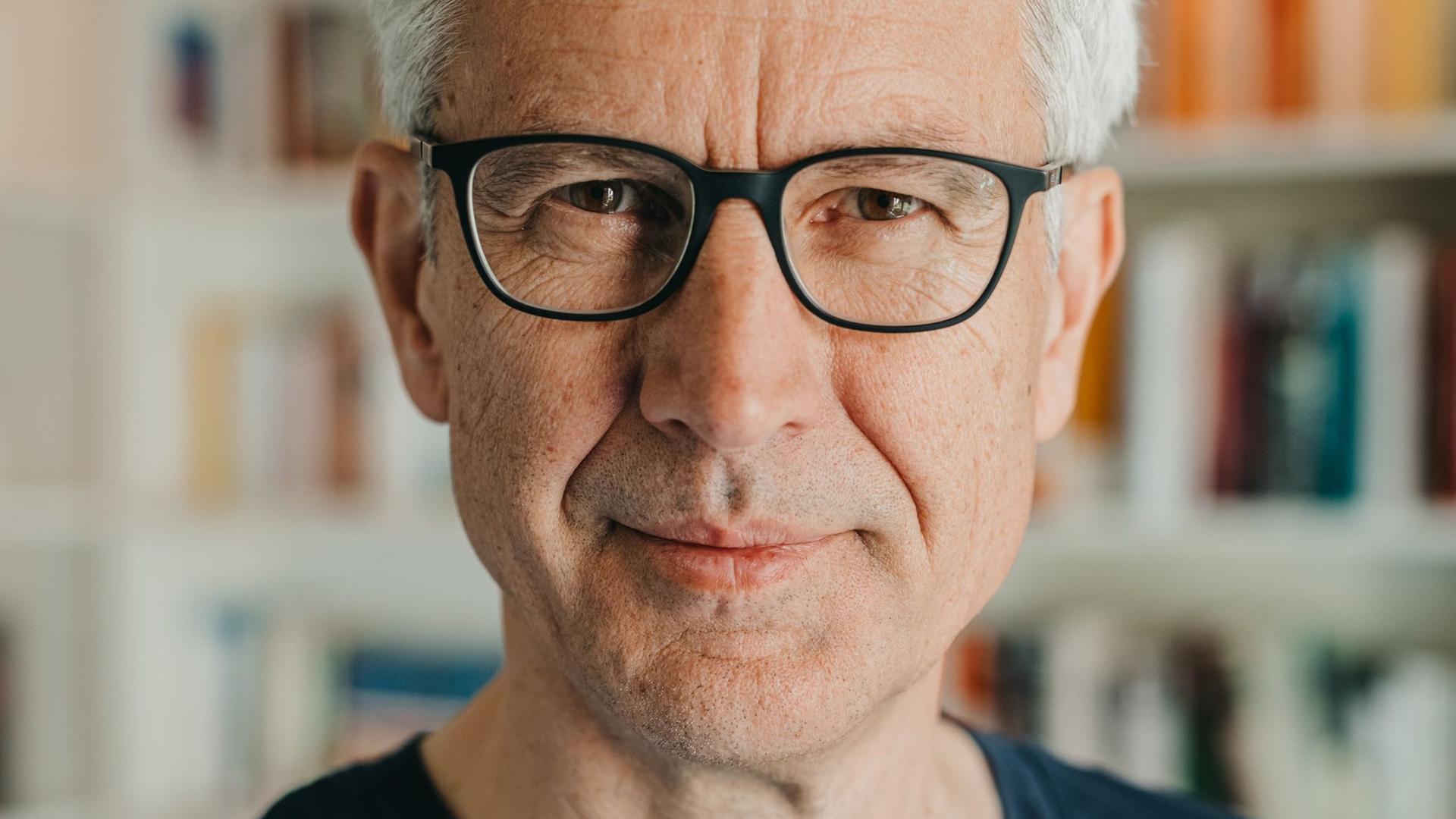 Ein Mann mit vollem, grauem Haar blickt freundlich in die Kamera. Er steht vor einer Bücherwand, trägt eine Brille mit schwarzem Rand und ein schwarzes T-Shirt. Ein Mann mit vollem, grauem Haar blickt freundlich in die Kamera. Er steht vor einer Bücherwand, trägt eine Brille mit schwarzem Rand und ein schwarzes T-Shirt.