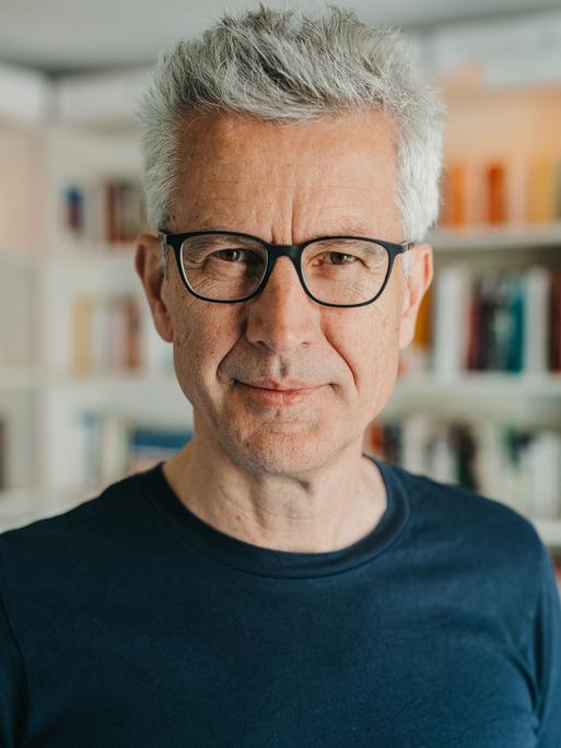 Ein Mann mit vollem, grauem Haar blickt freundlich in die Kamera. Er steht vor einer Bücherwand, trägt eine Brille mit schwarzem Rand und ein schwarzes T-Shirt.