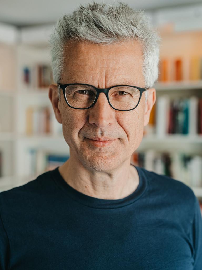 Ein Mann mit vollem, grauem Haar blickt freundlich in die Kamera. Er steht vor einer Bücherwand, trägt eine Brille mit schwarzem Rand und ein schwarzes T-Shirt.