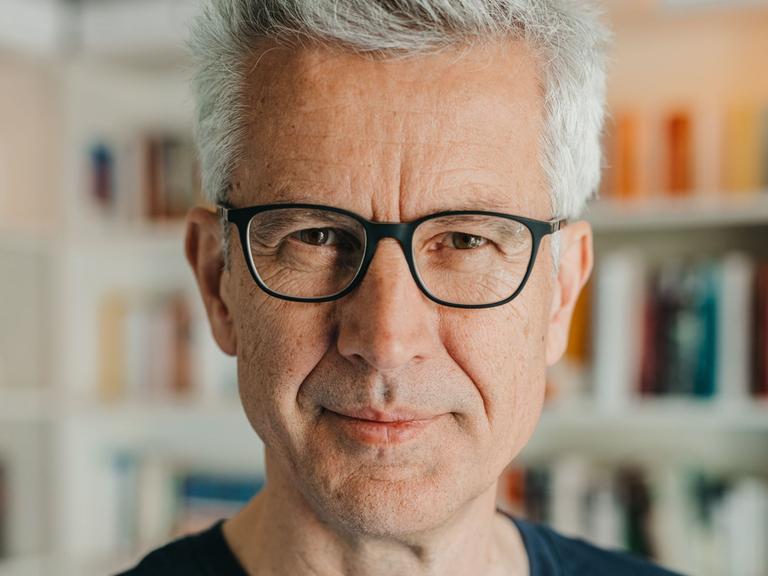 Ein Mann mit vollem, grauem Haar blickt freundlich in die Kamera. Er steht vor einer Bücherwand, trägt eine Brille mit schwarzem Rand und ein schwarzes T-Shirt.