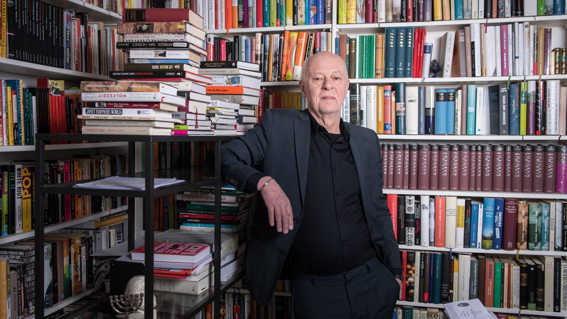 Schriftsteller und Verleger Klaus Bittermann in den Räumen seines Verlags Edition Tiamat in Berlin Kreuzberg. Er steht in einem grauen Anzug vor einem vollen Bücherregal, Buchstapel liegen um ihn herum. Schriftsteller und Verleger Klaus Bittermann in den Räumen seines Verlags Edition Tiamat in Berlin Kreuzberg. Er steht in einem grauen Anzug vor einem vollen Bücherregal, Buchstapel liegen um ihn herum.